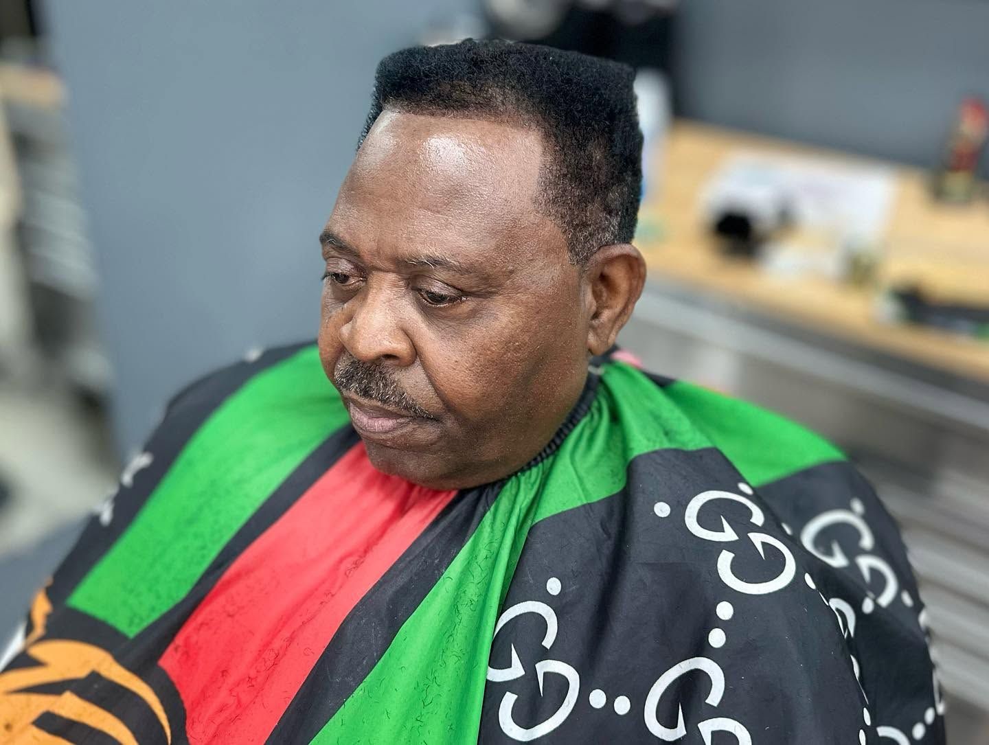 Man seated in a black, green, and red barber cape, looking down in a salon chair