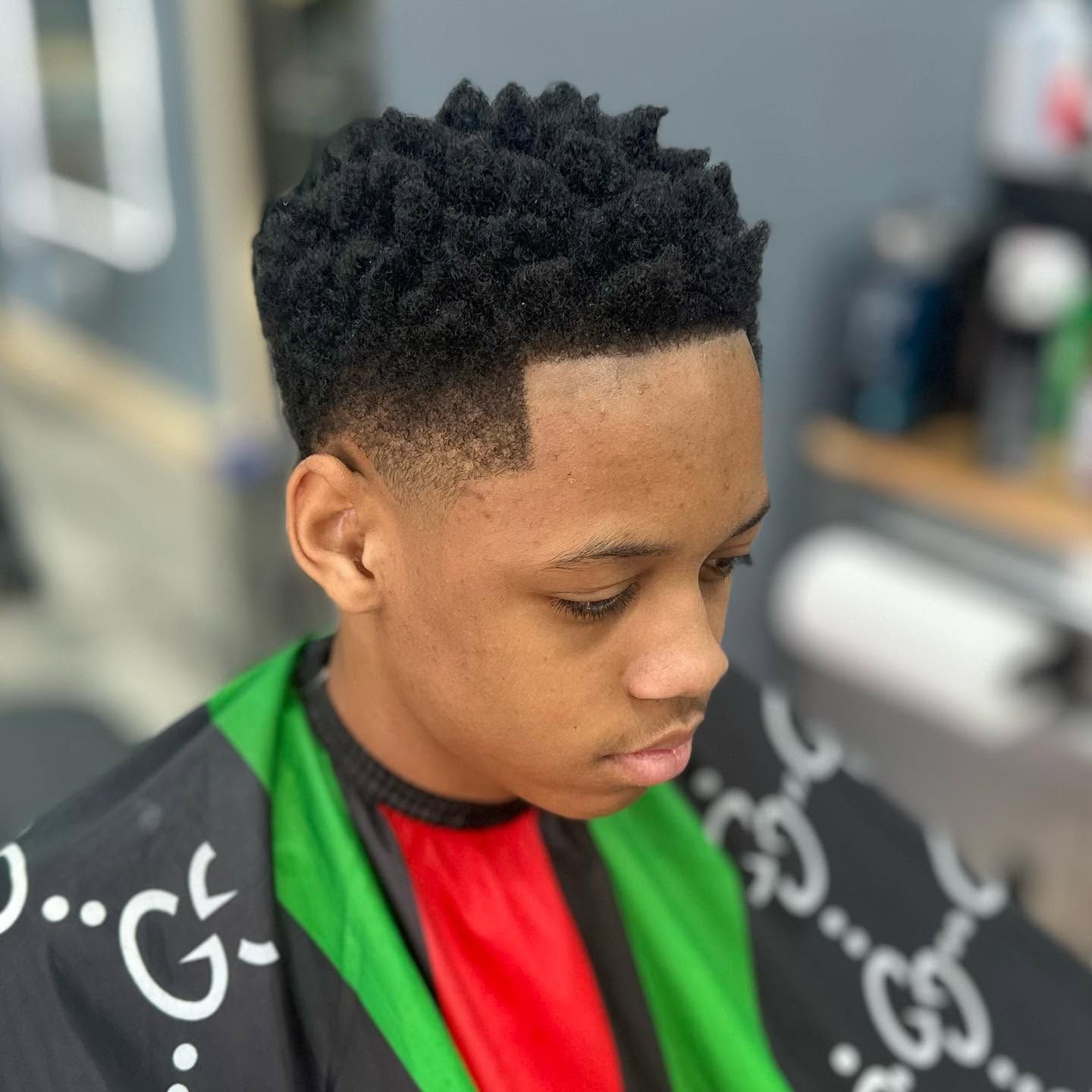 Person with short curly hair wearing a red and green barber cape, looking down in a salon chair.