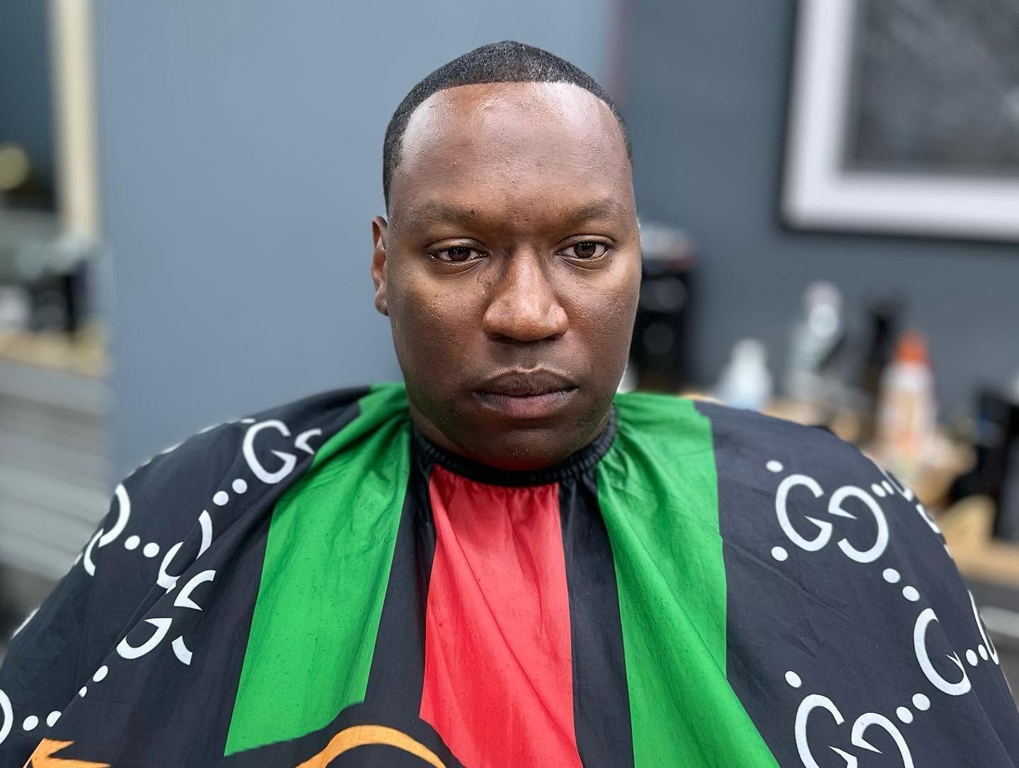 Man seated indoors wearing a black, green, and red patterned barber cape, looking at the camera.