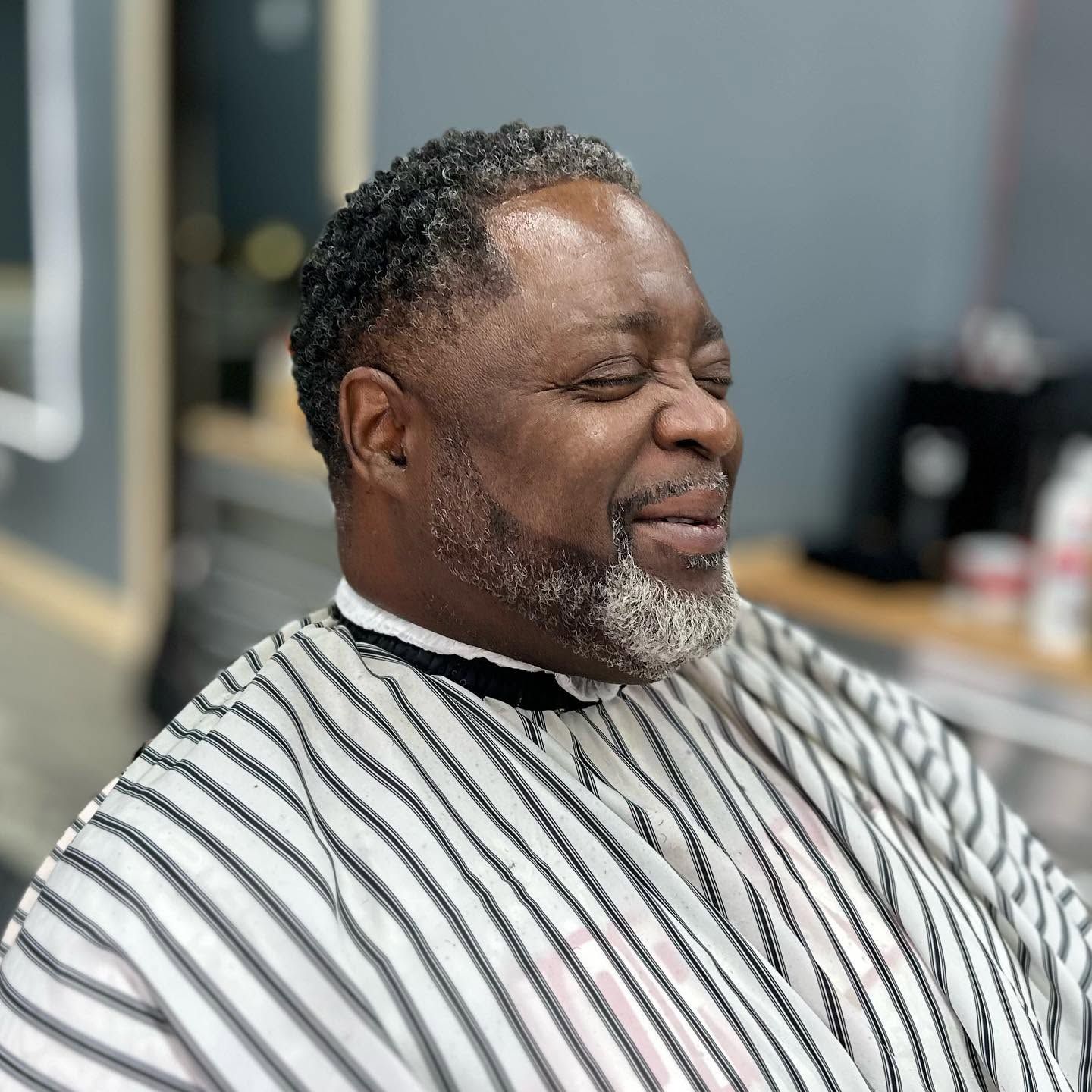 Man sitting in a barber chair wearing a striped cape, smiling in a barbershop