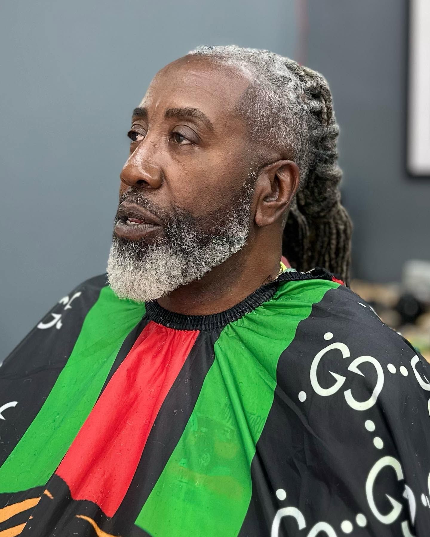 Man with gray beard wearing a black, red, and green barber cape, seated indoors looking to the side