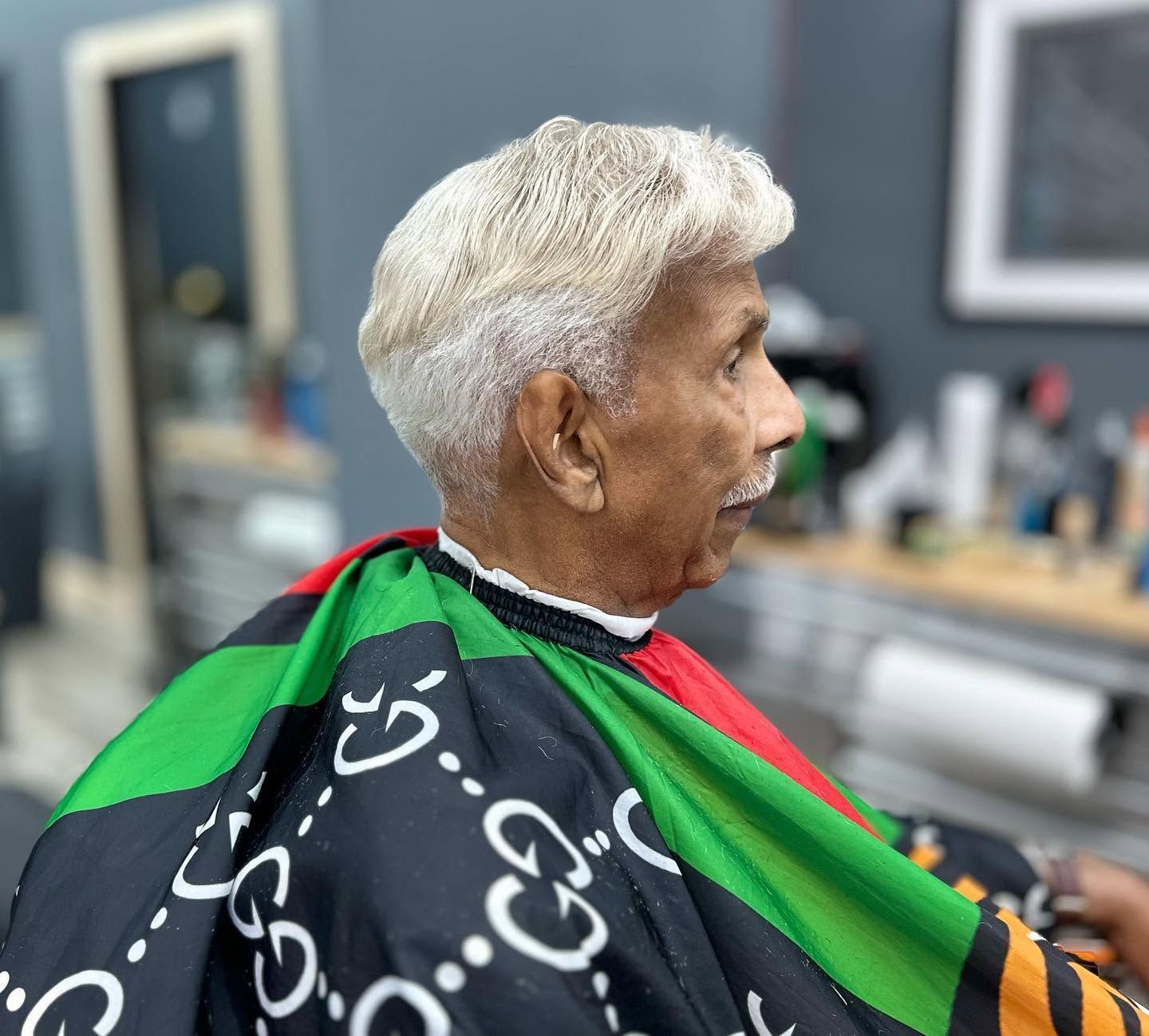 Older person with short white hair seated in a salon chair, wearing a black, green, and red cape.