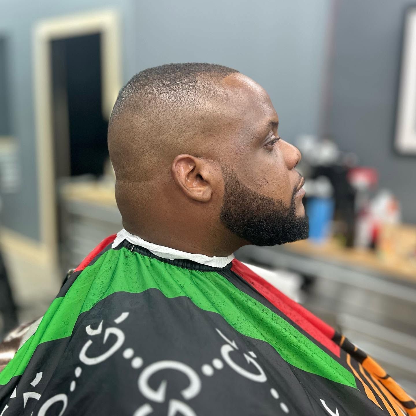 Side-profile of a man with a beard getting a haircut in a barbershop, wearing a green and black cape.