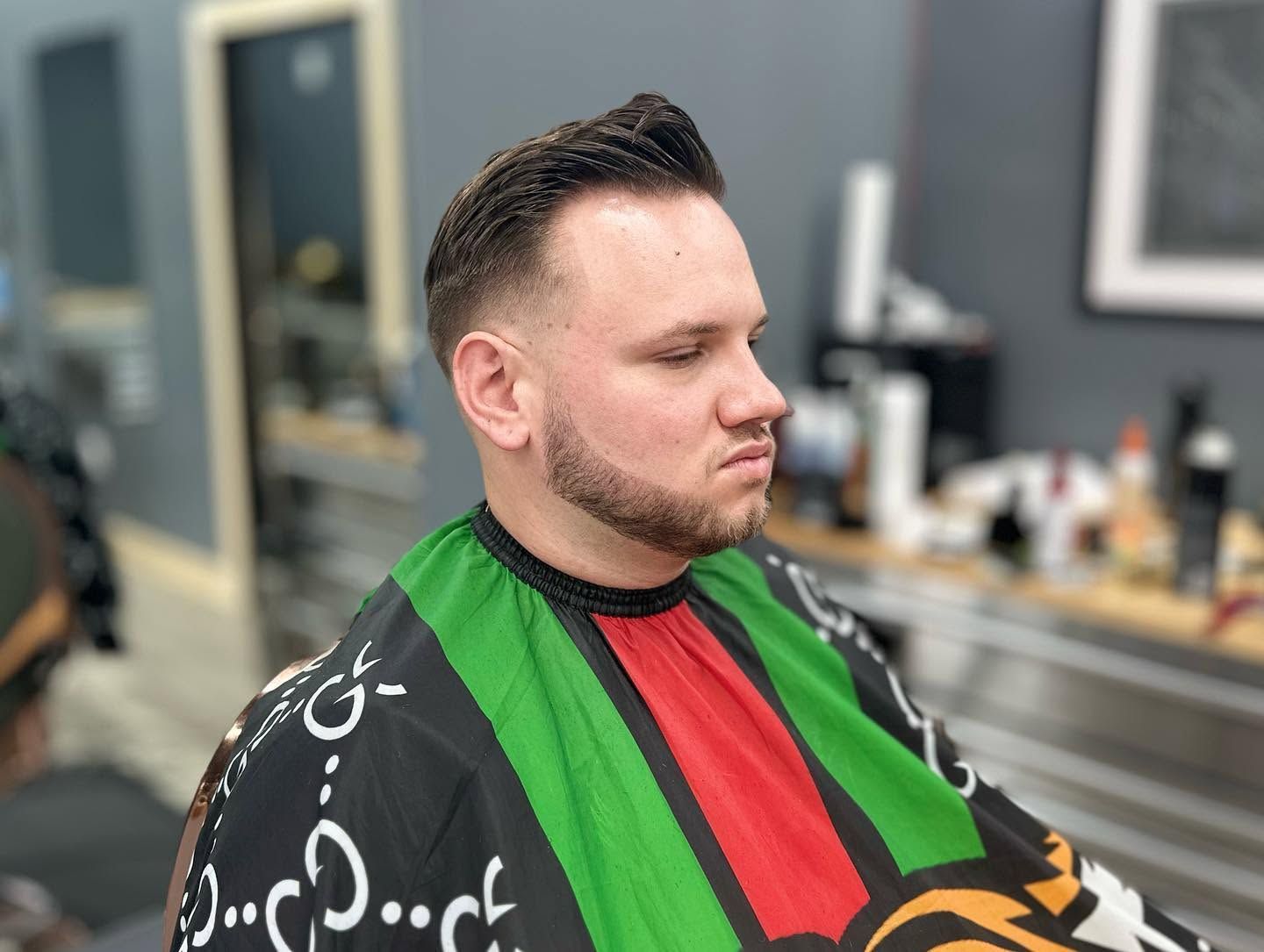Man in barber cape with green, red, and black stripes, sitting in a salon chair during a haircut