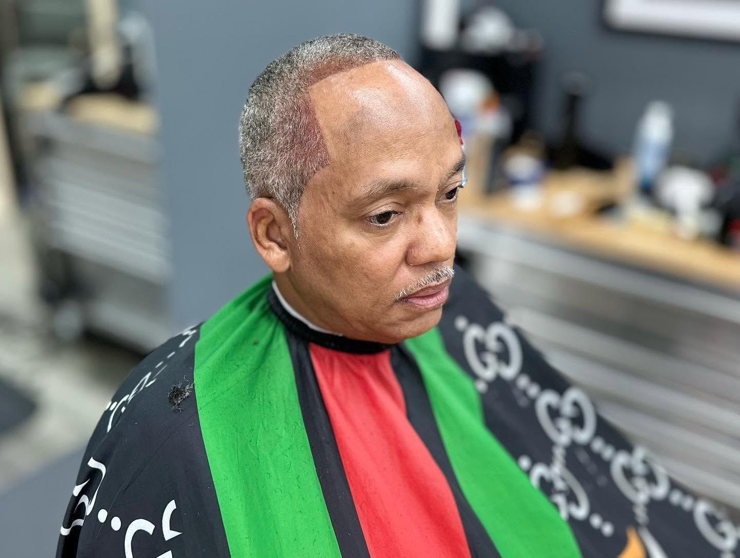 Man in a black barber cape with green and red stripes, seated in a barbershop chair