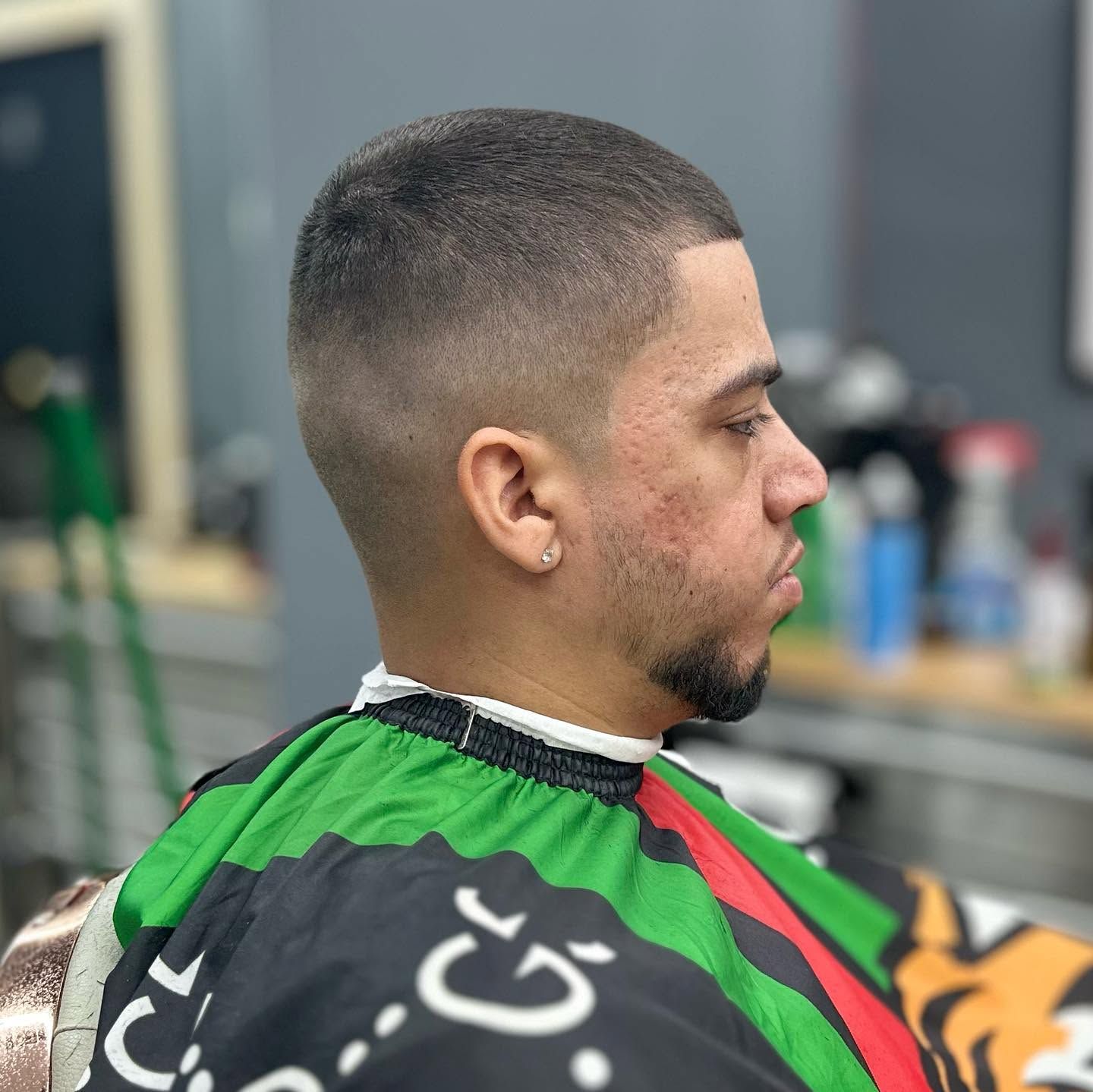 Side-profile of a man with a short fade haircut wearing a green barber cape in a barbershop.
