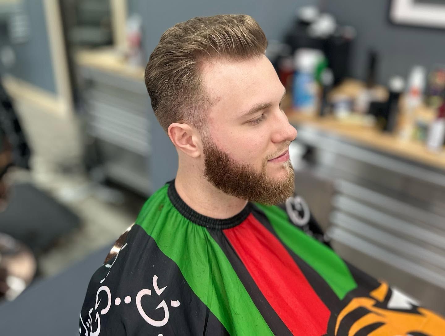 Man in a black, green, and red barber cape seated in a barbershop, turned slightly to the side.