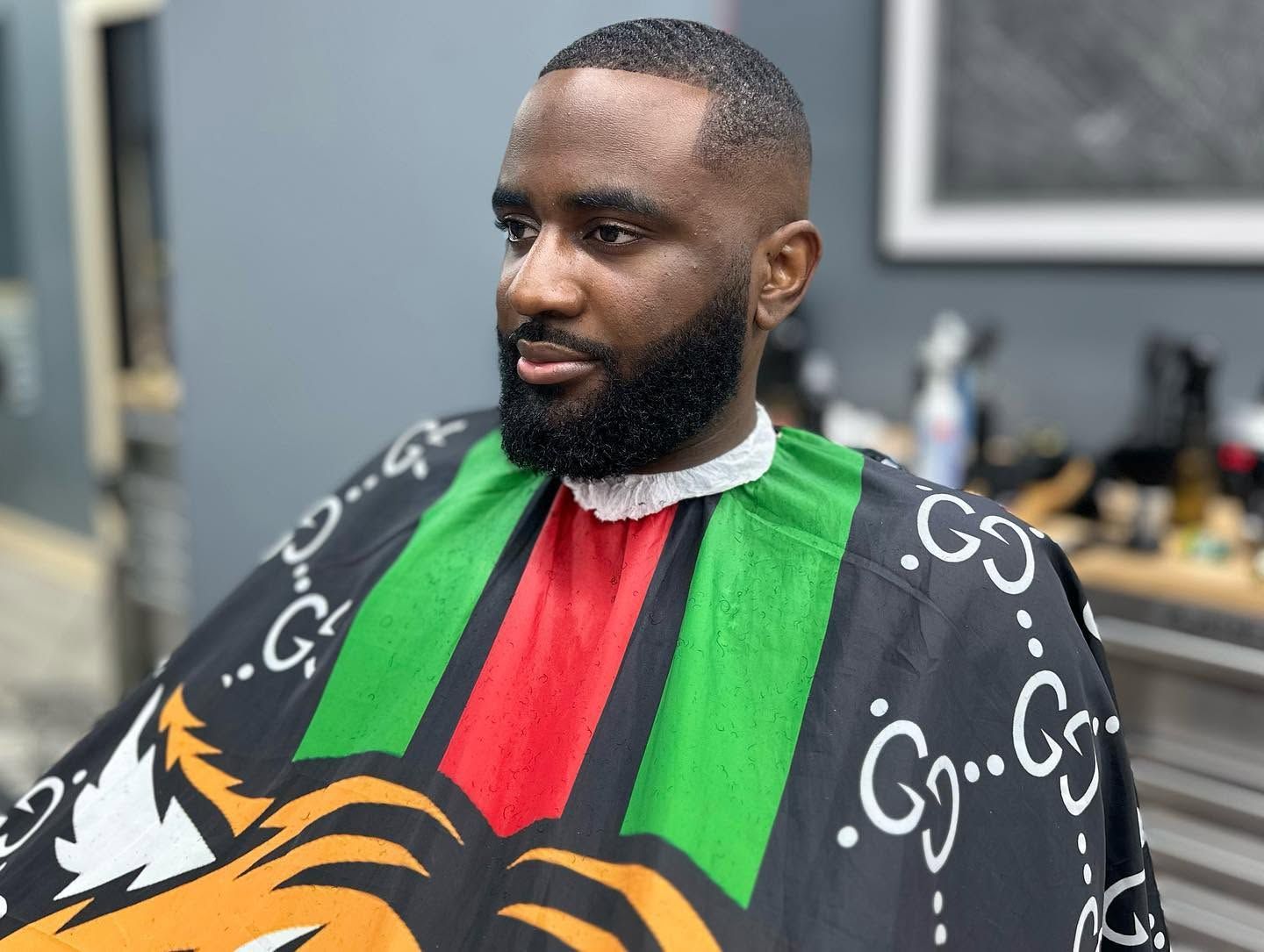 Man in a barber cape with green, red, and black stripes, seated indoors with a neatly trimmed beard.
