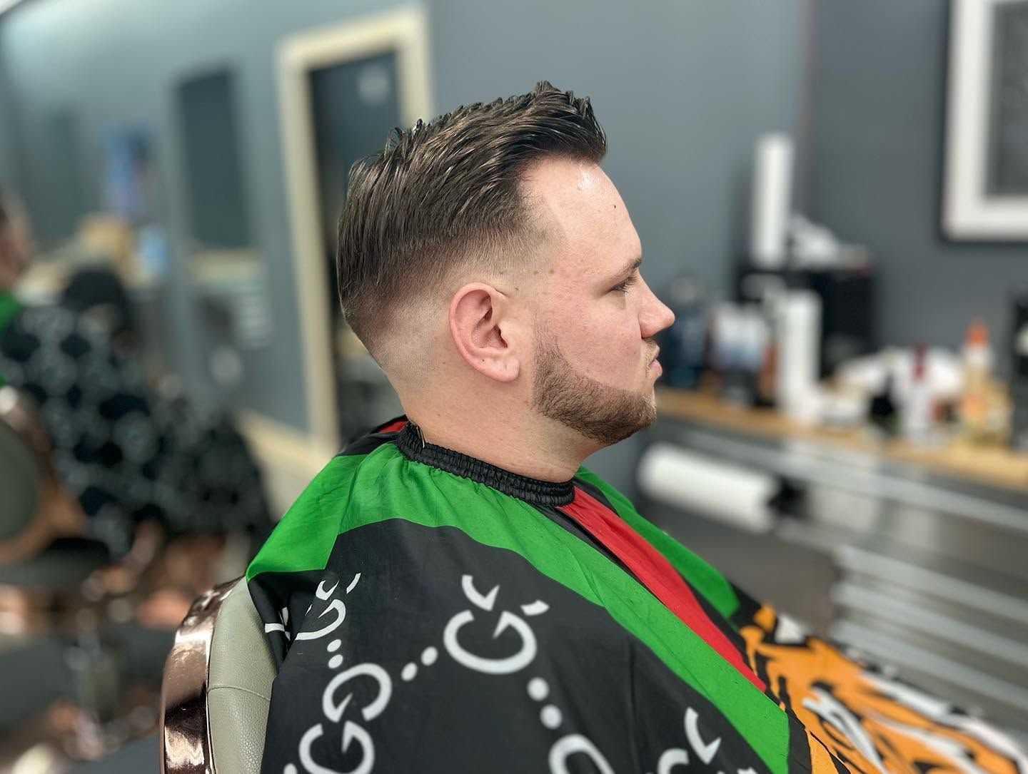 Man with a fade haircut in a barber chair, wearing a black-and-green cape, viewed in profile.