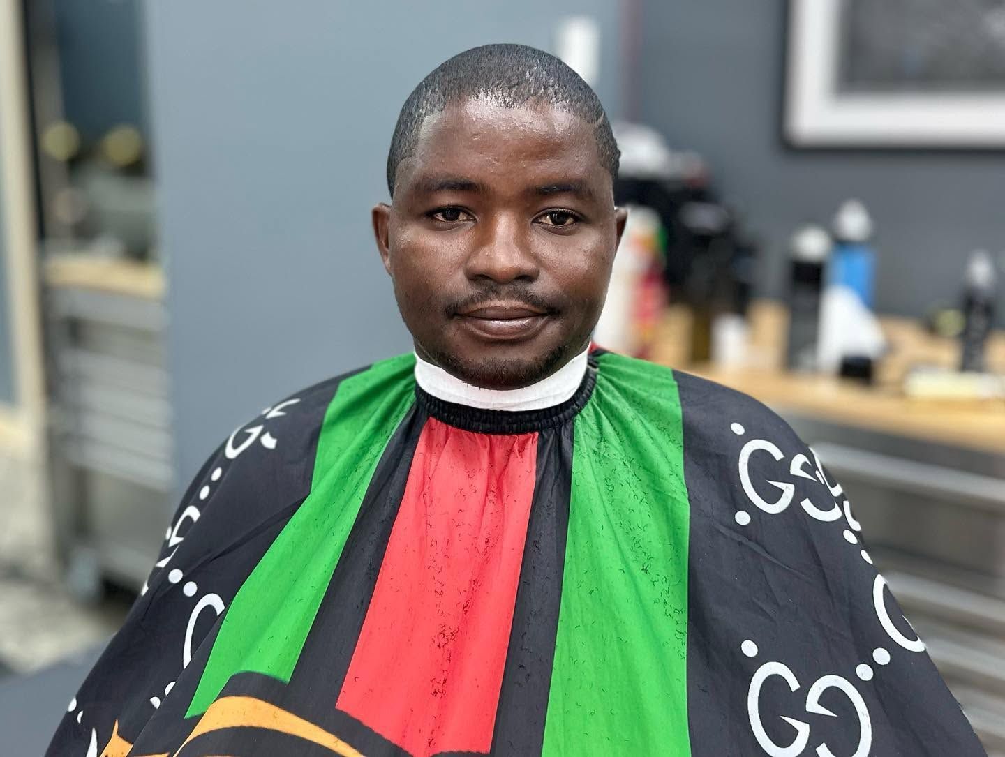 Person seated in a barber chair wearing a black, green, and red barber cape, looking at the camera.