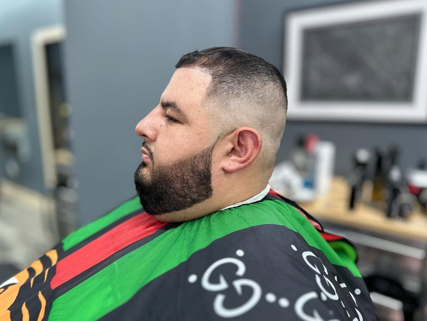 Man seated in a barber chair wearing a black cape with green and red stripes, in a barbershop.