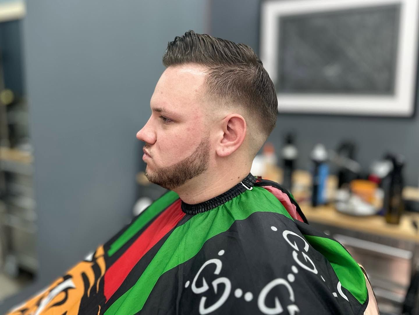 Man seated in a barber chair wearing a green and red cape, getting a haircut in a barbershop
