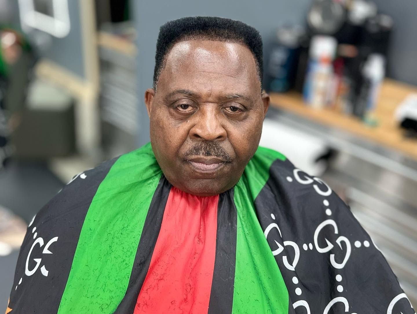 Person in a green, red, and black barber cape sitting in a salon chair with a neutral expression