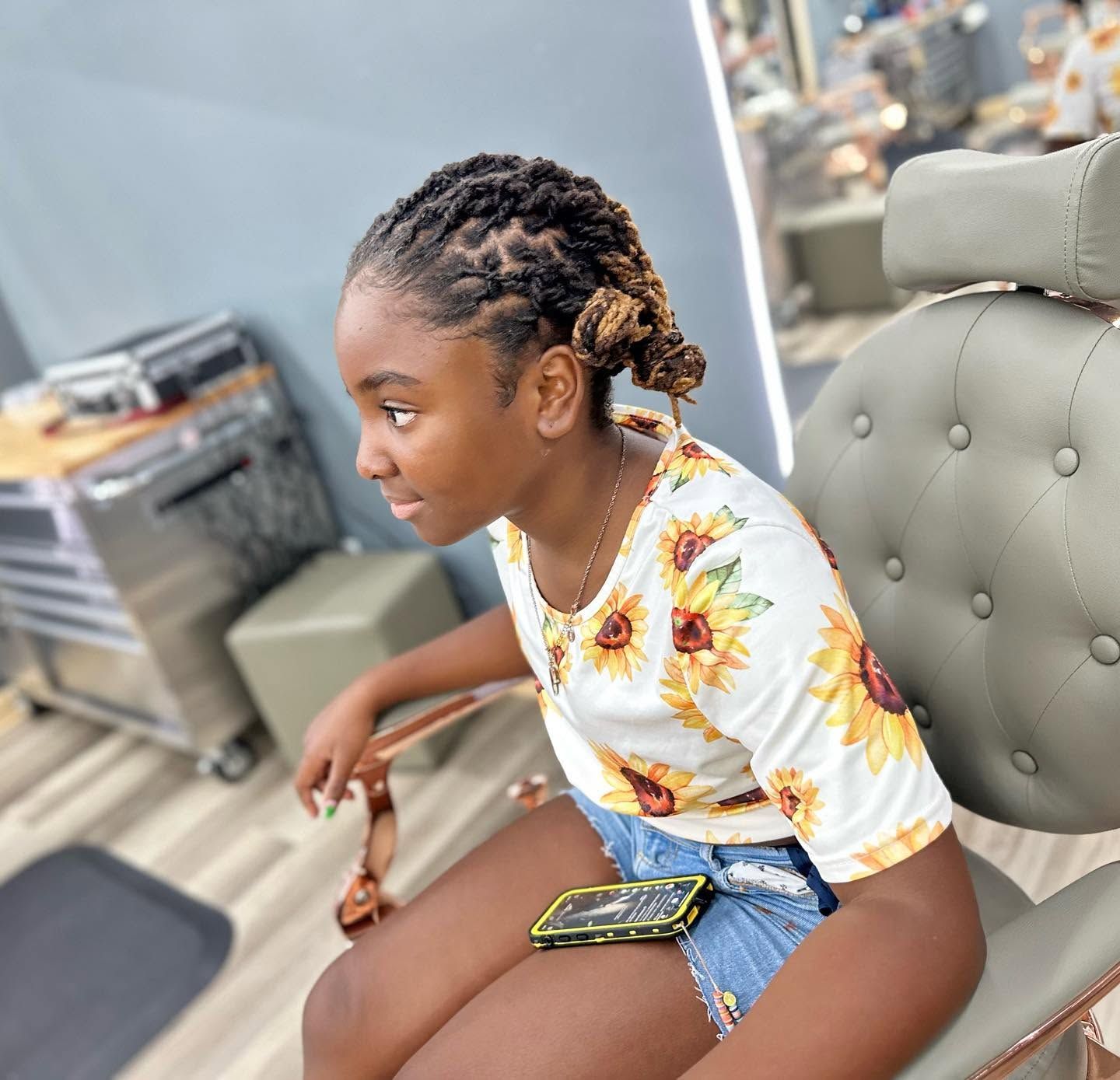 Woman seated in a salon chair, wearing a sunflower top and denim shorts, looking left while holding a phone.