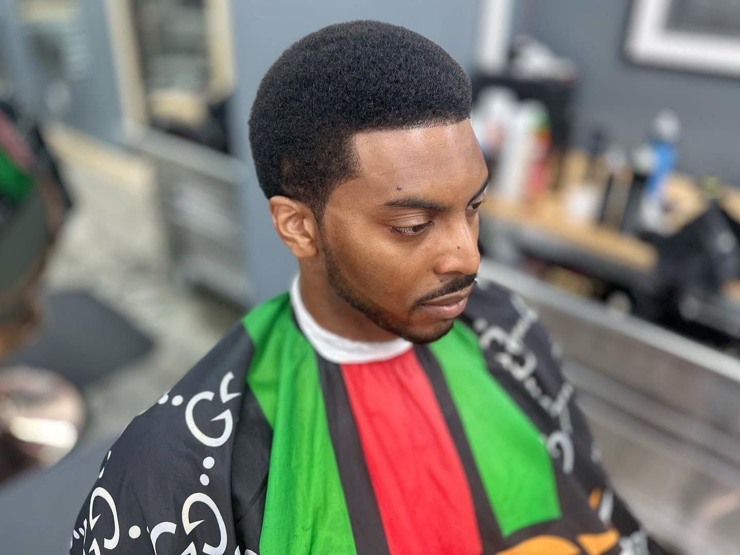 Man wearing a green, red, and black barber cape, seated in a salon chair with a blurred background.