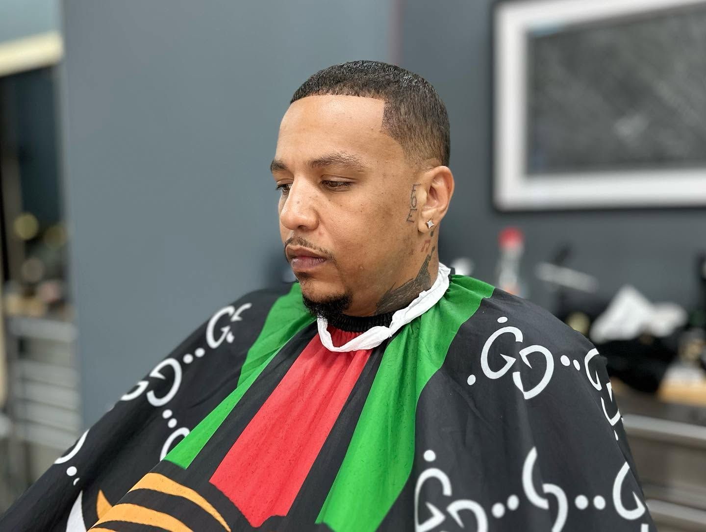 Man sitting in a barber chair, wearing a black cape with green and red stripes, looking down.