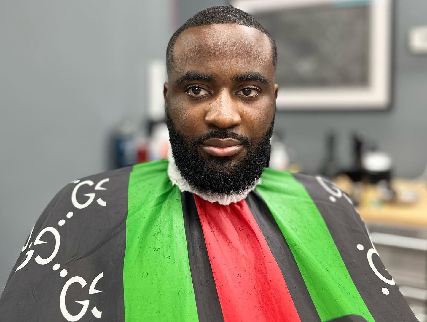 Person in a black, green, and red barber cape seated indoors, looking at the camera.