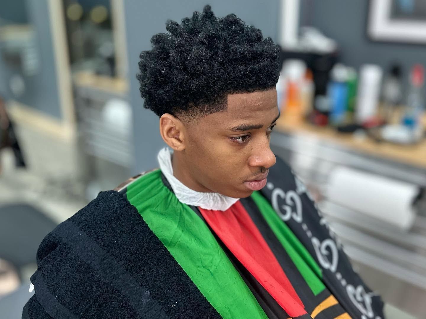 Person in a barber chair wearing a black, green, and red cape, looking down in a barbershop.
