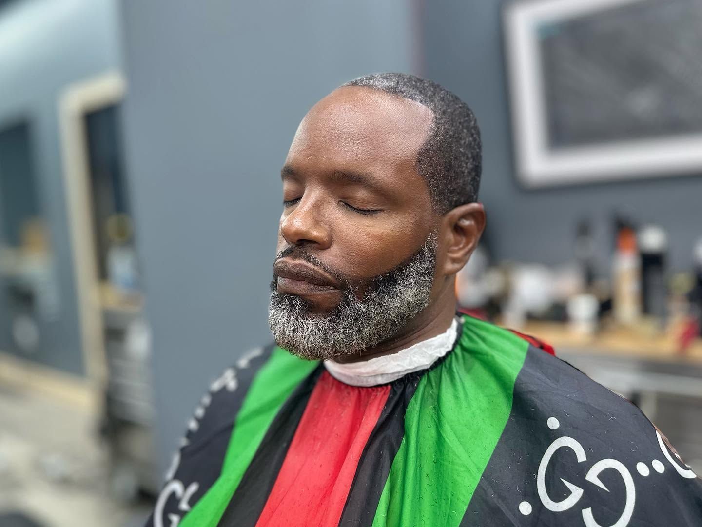 Person in a barber chair with eyes closed, wearing a black, red, and green cape in a salon.