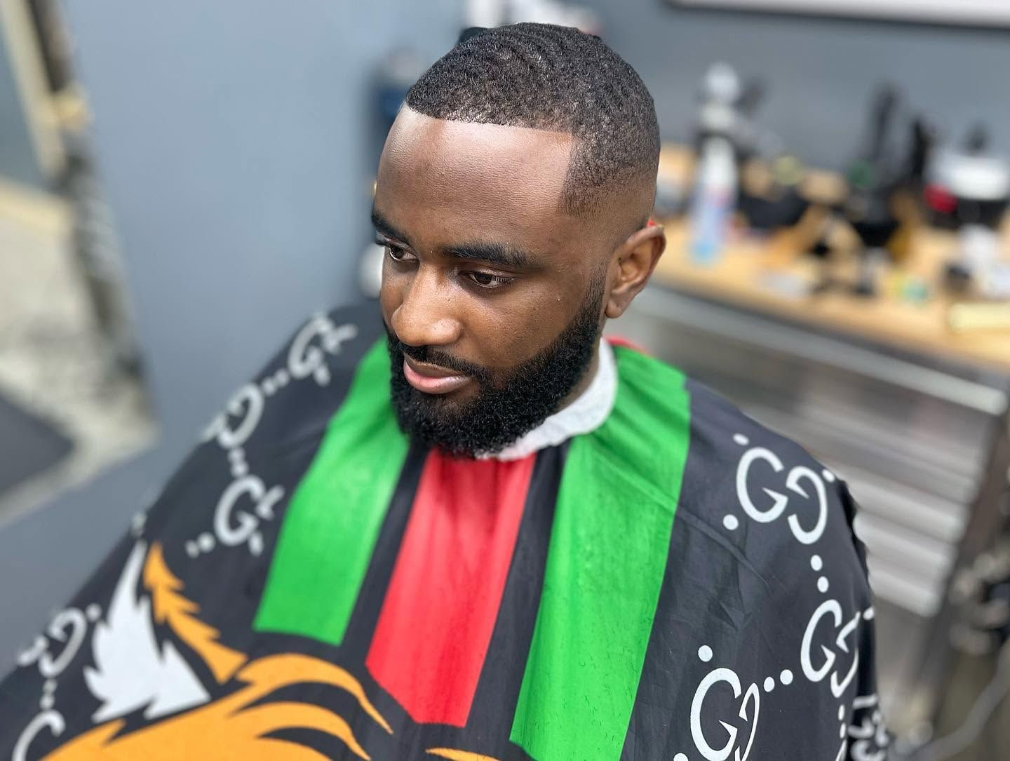 Man in a black, red, and green striped barber cape, looking down in a barbershop chair.