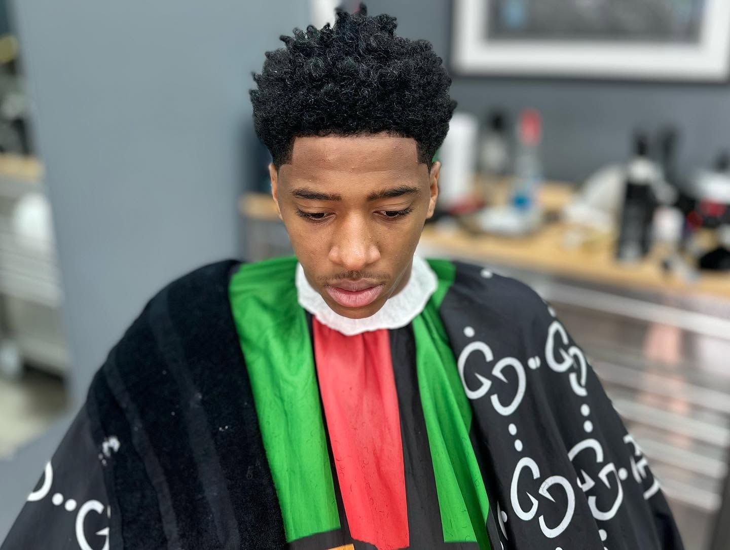 Person in a barber cape with green and red stripes, seated in a salon and looking down.