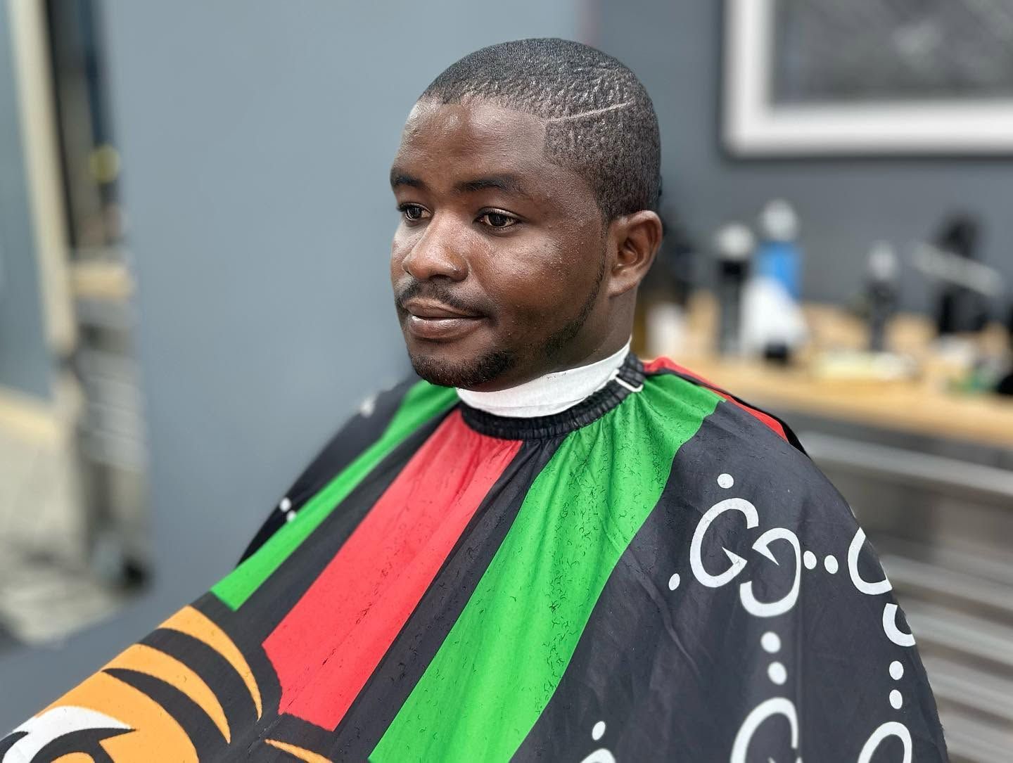 Man sitting in a barber chair wearing a black cape with green and red stripes, looking to the side.