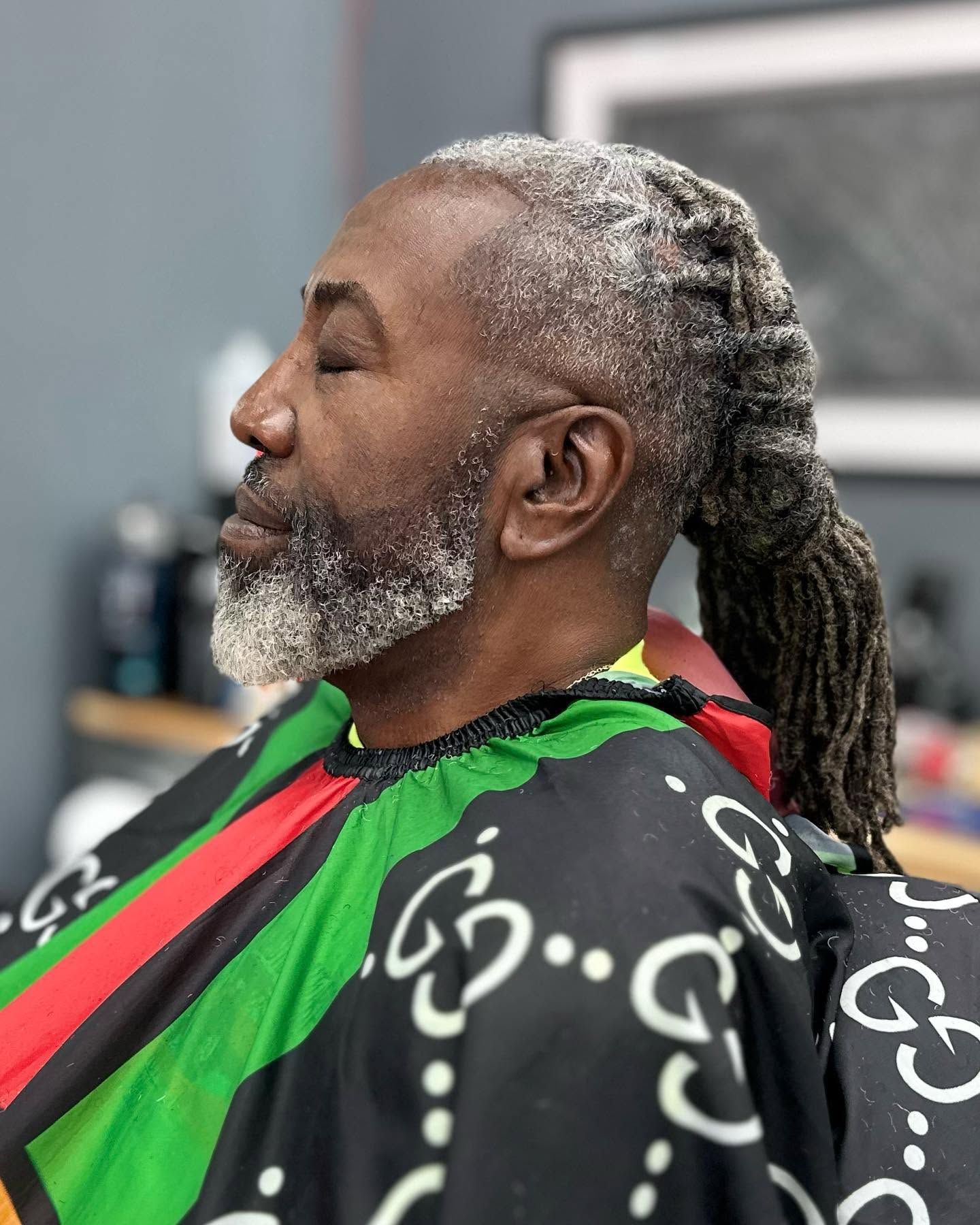 Profile view of a man with gray beard and locs wearing a black-and-red barber cape