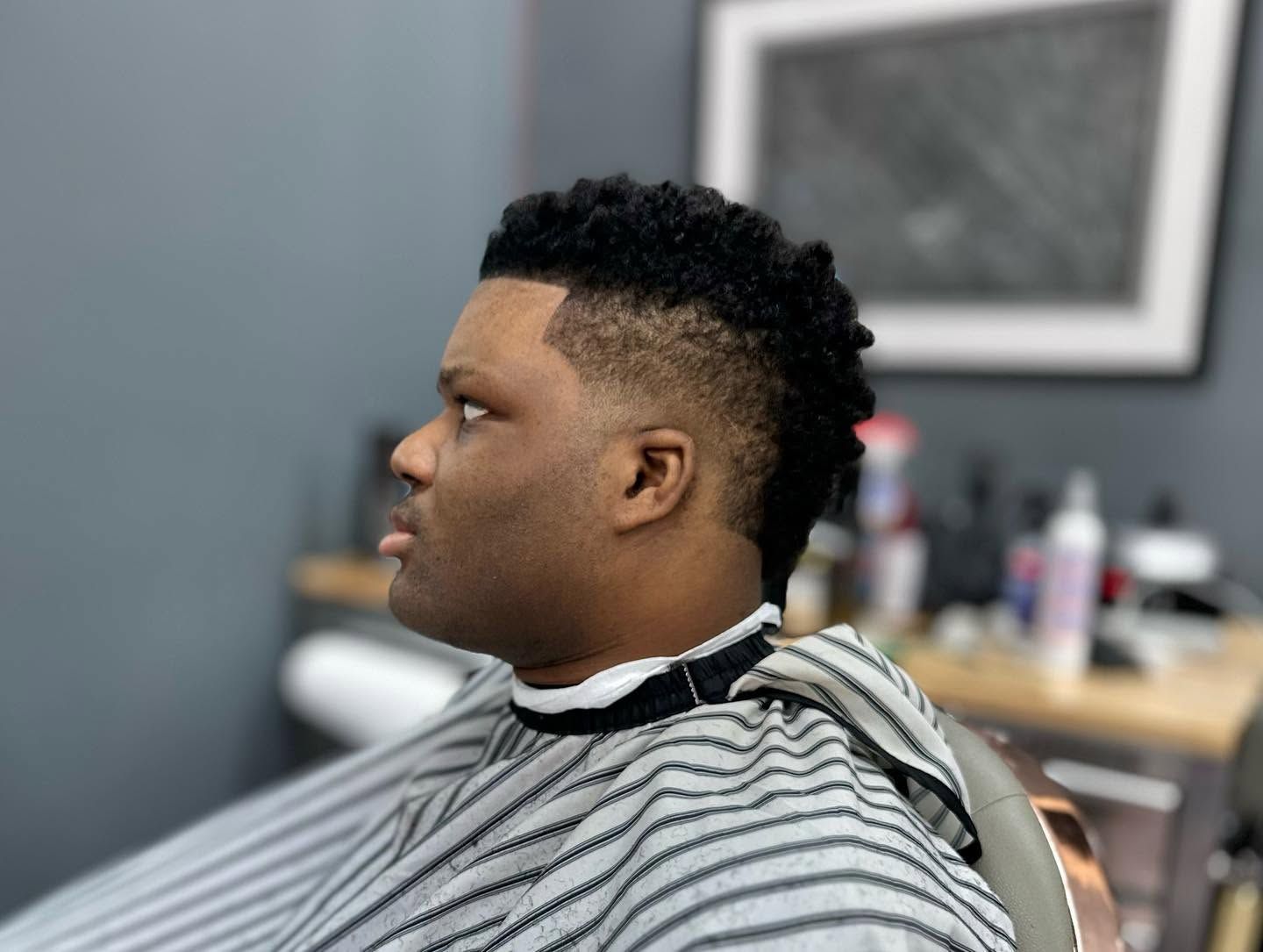 Person with a short curly haircut seated in a barber chair, wearing a striped cape in a salon.