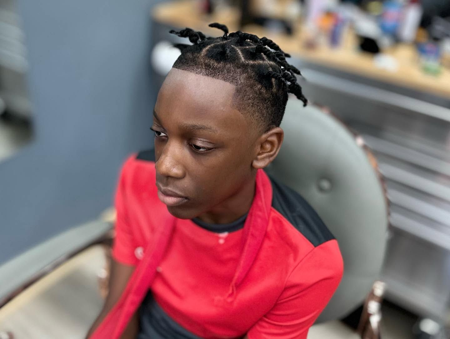 Person in a red shirt sitting in a salon chair, looking down with braided hair.