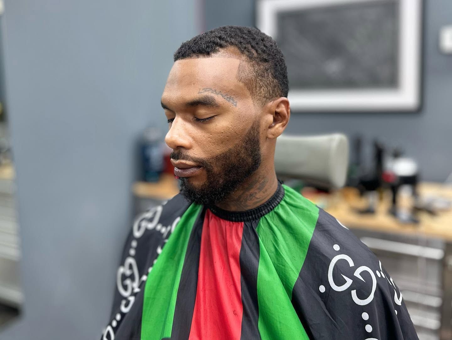 Man seated in a barber chair wearing a green, red, and black cape, with a short beard and closed eyes
