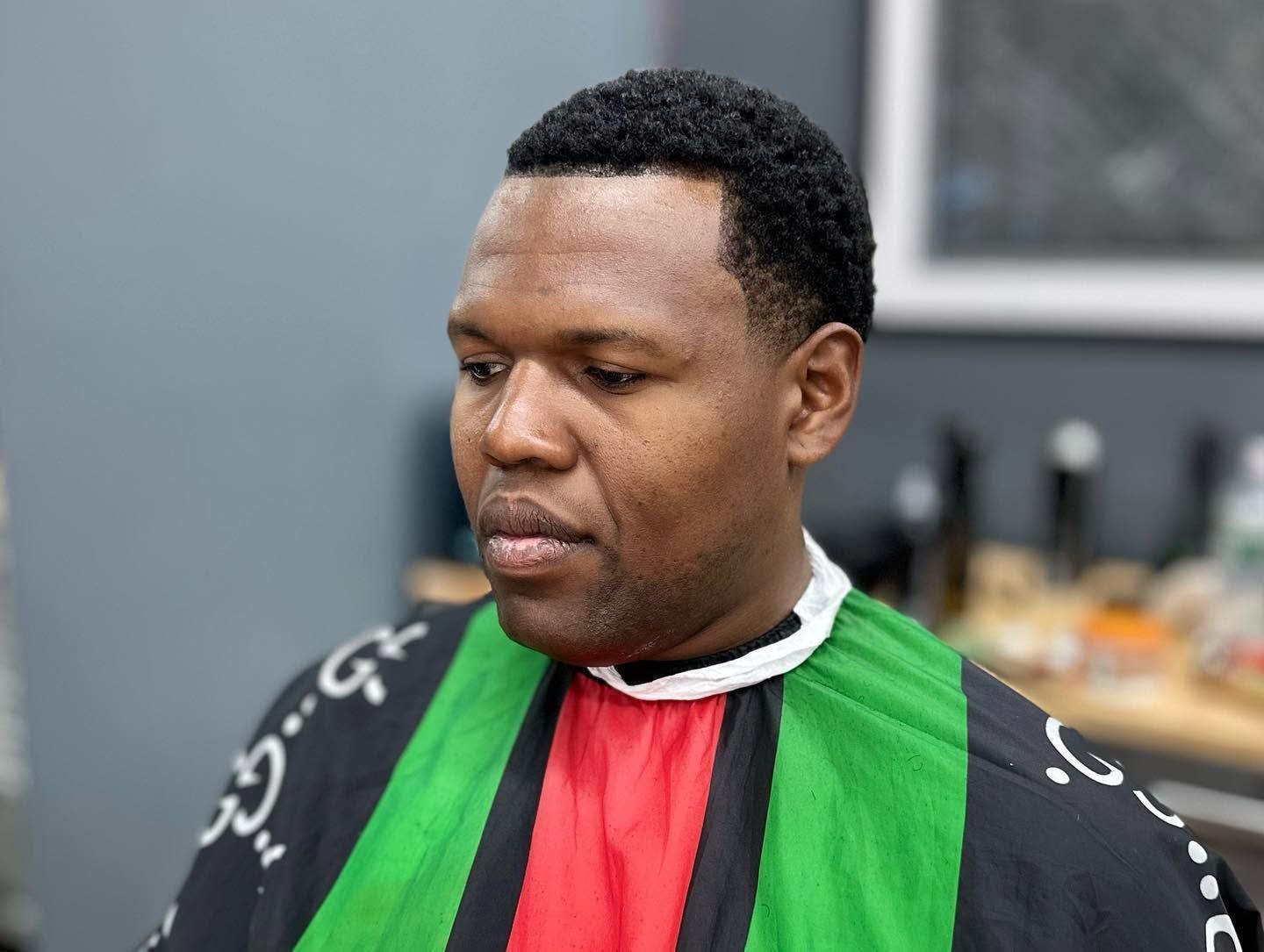 Person wearing a black, green, and red barber cape, seated in a salon, looking down thoughtfully