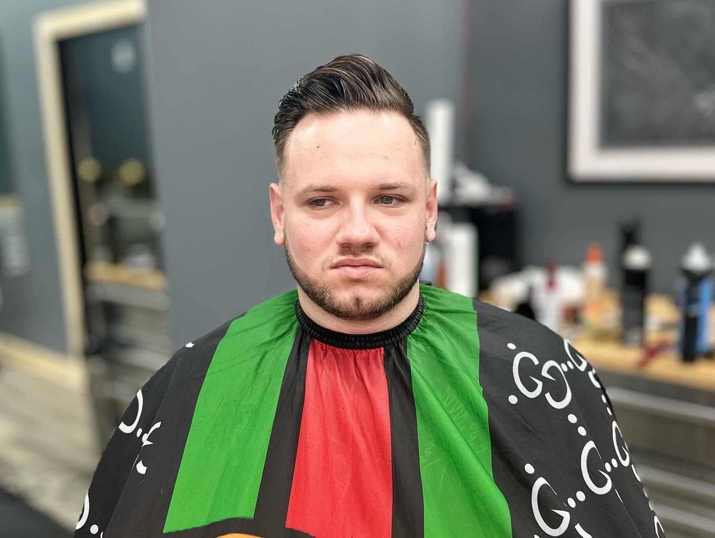 Person seated in a barber chair wearing a black cape with green and red stripes, in a salon.