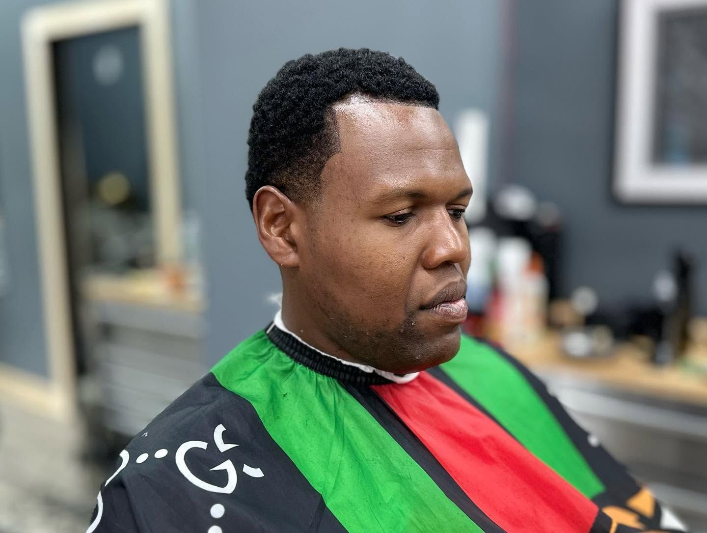 Person in a barber chair wearing a green, red, and black striped cape, looking down in a salon.