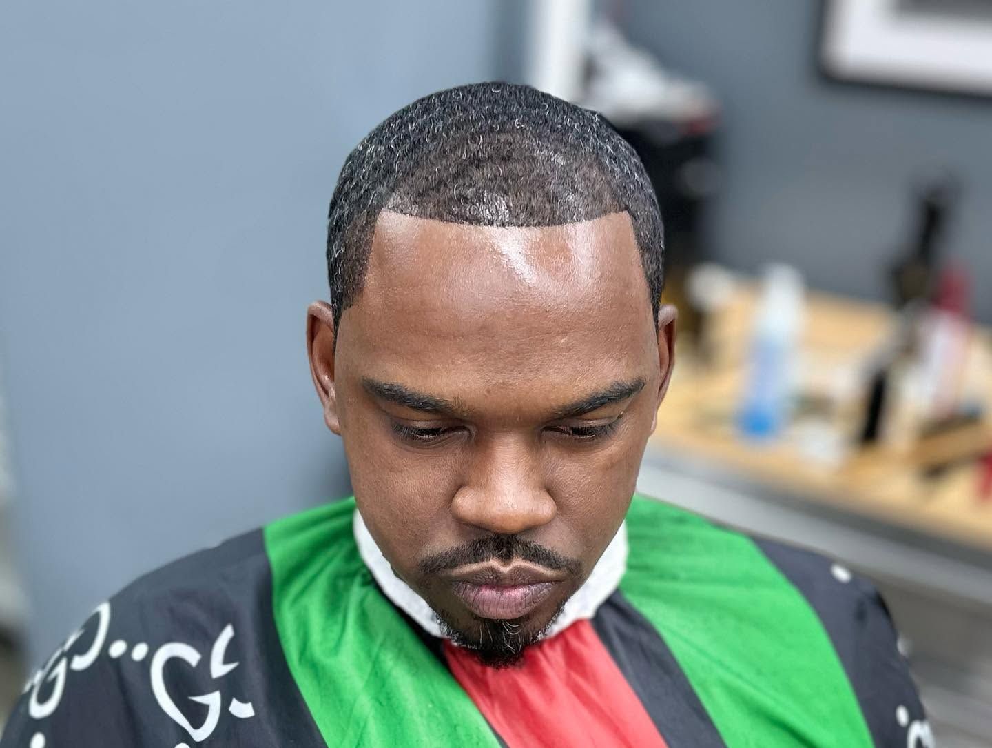 Man with a fresh haircut and trimmed beard, looking down in a barber chair with a red, green, and black cape.
