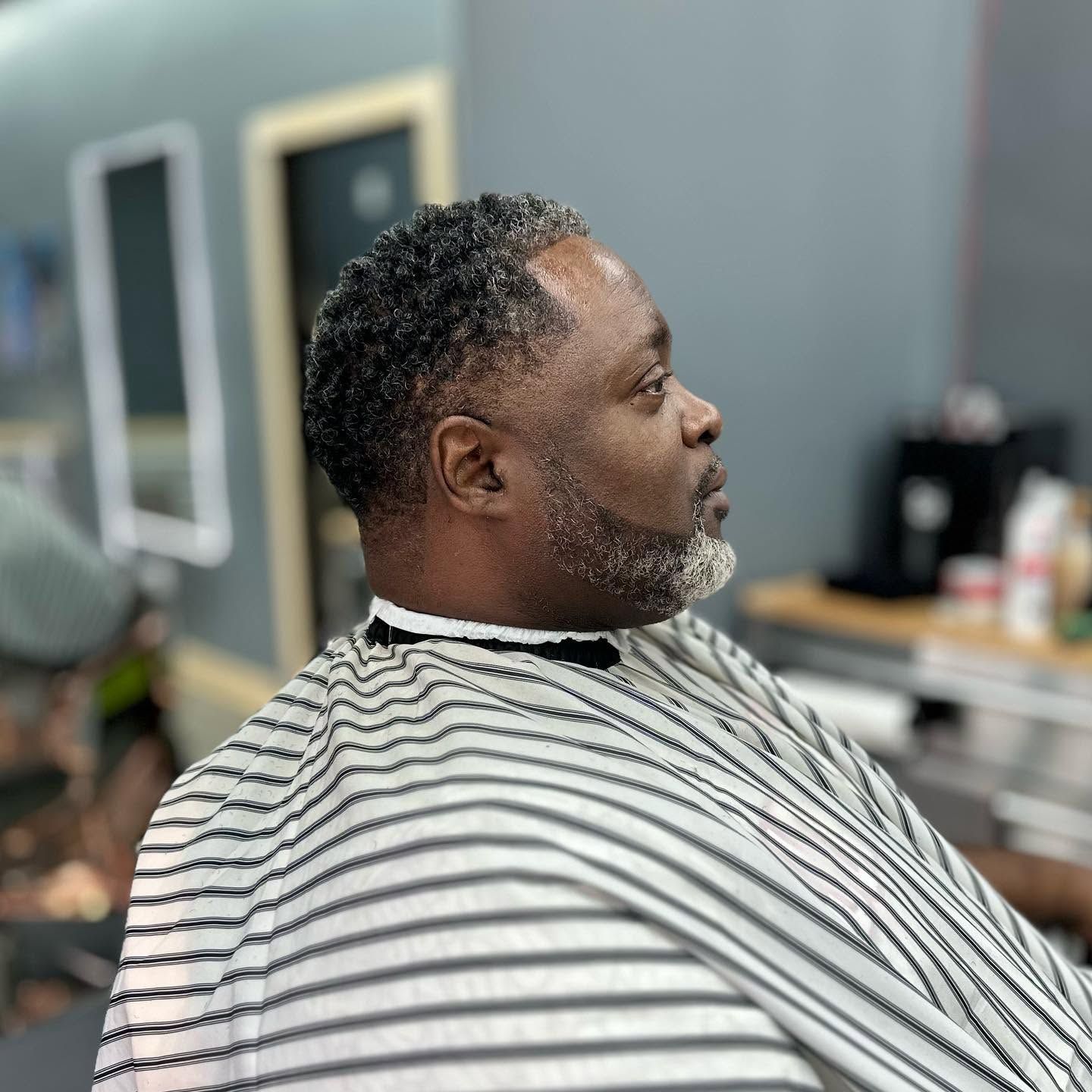 Side profile of a man in a barber chair, wearing a striped cape in a modern barbershop.