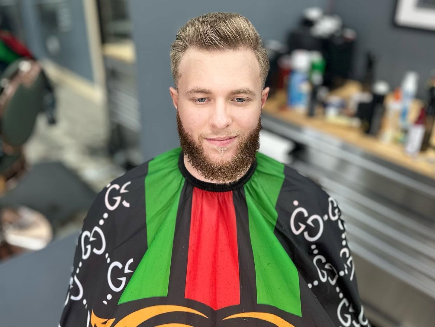 Man seated in a barbershop chair wearing a black cape with green and red stripes, looking down with a slight smile