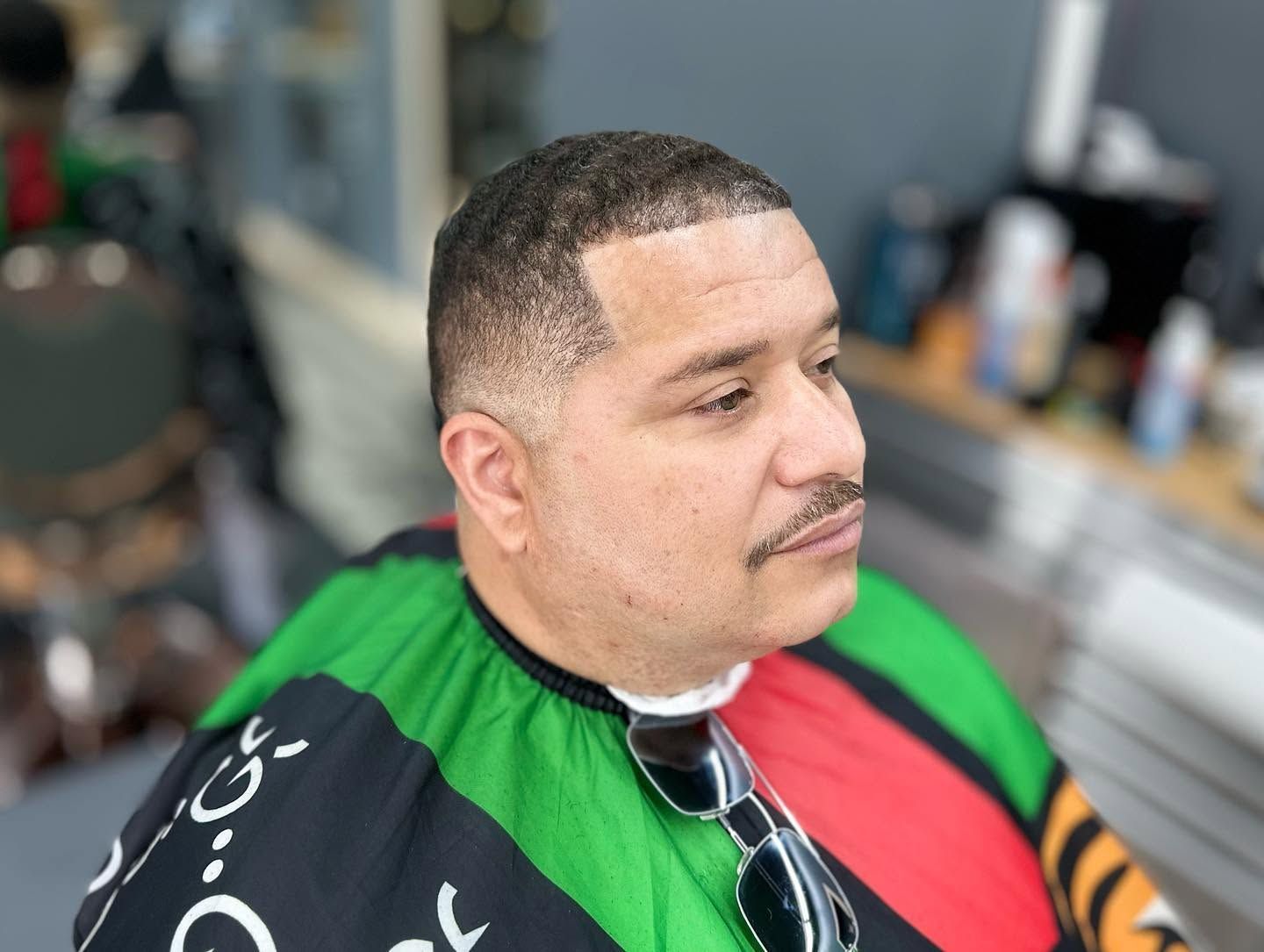 Man in a barber chair wearing a green, red, and black cape, with clippers visible nearby.