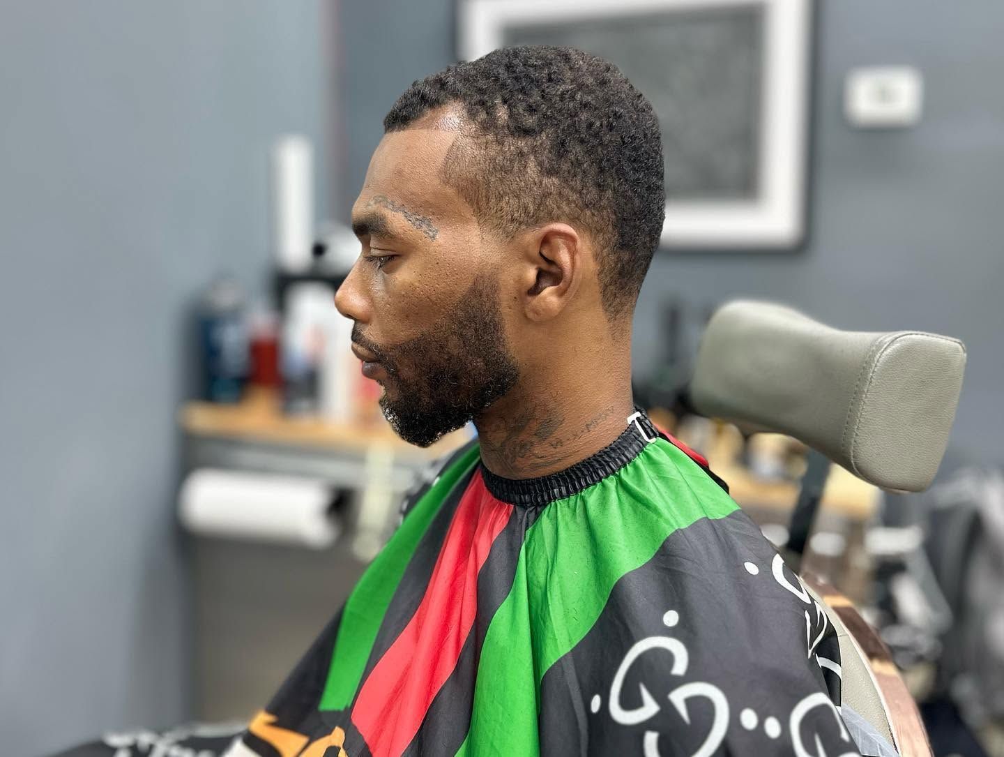 Profile view of a man getting a haircut in a barbershop, wearing a green, red, and black cape.