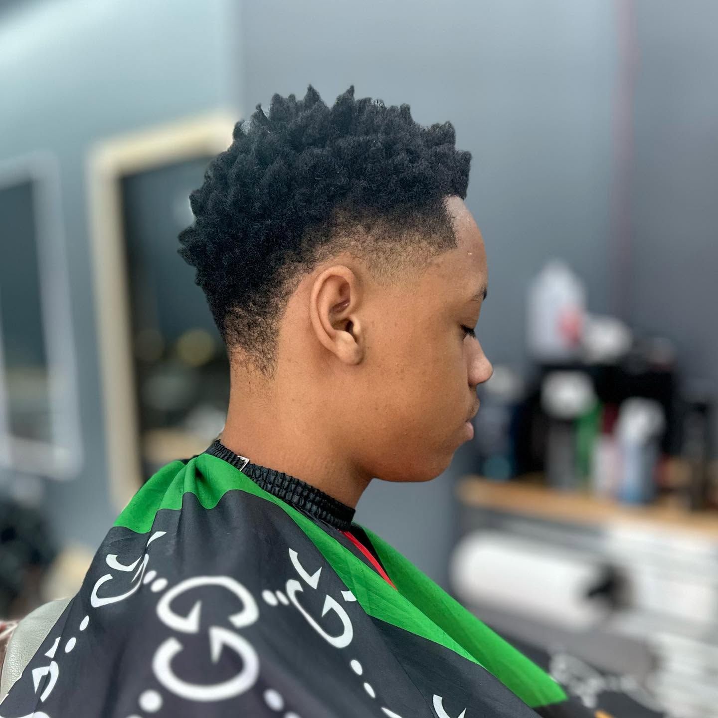 Person getting a haircut in a barbershop, wearing a black cape with green trim.