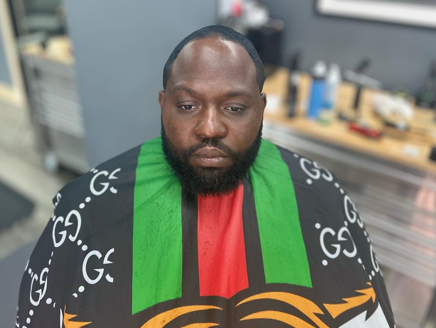 Man wearing a black, green, and red patterned jacket, seated indoors with a neutral expression.