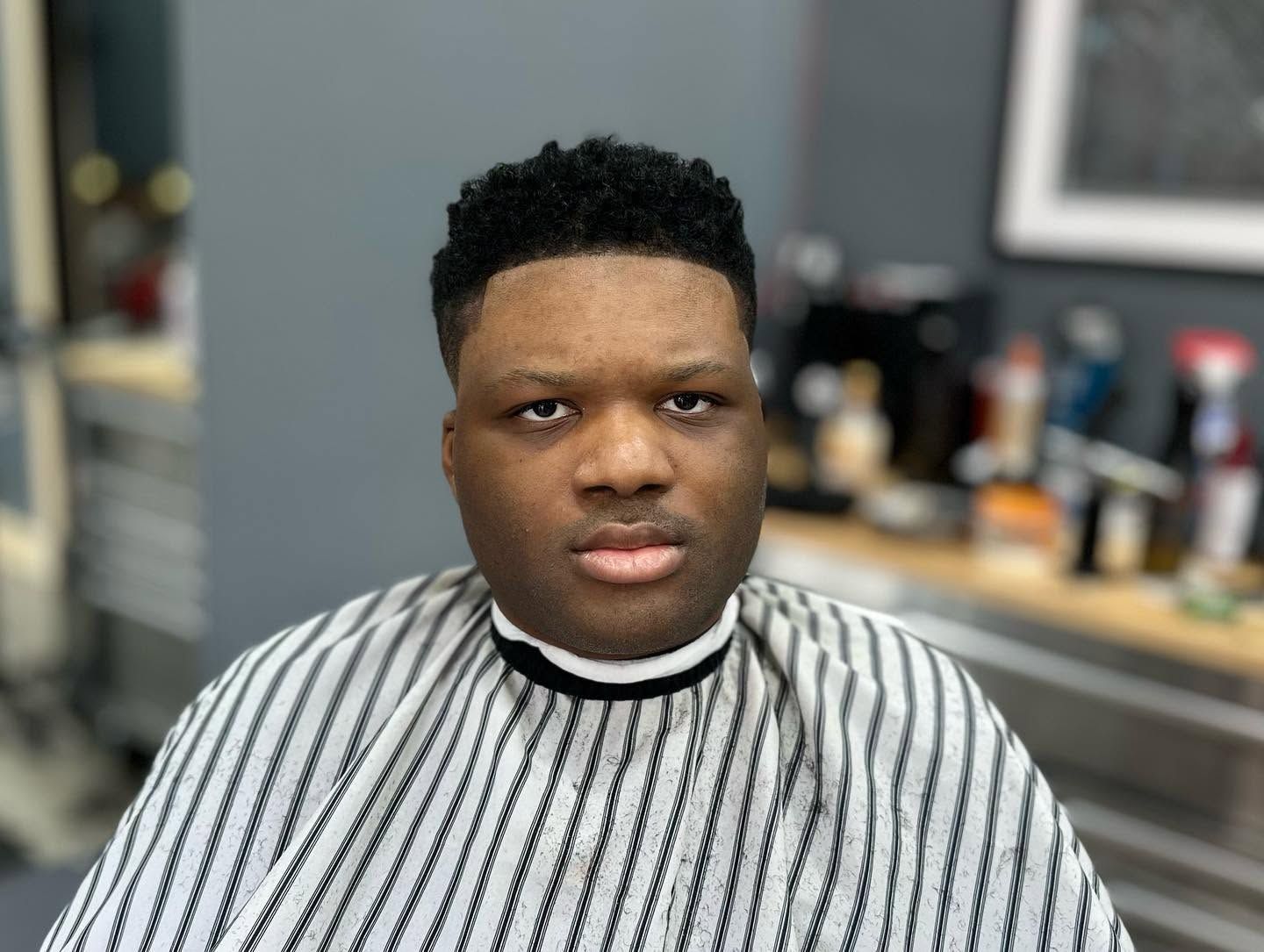 Person seated in a barber chair wearing a striped cape, looking at the camera in a salon.