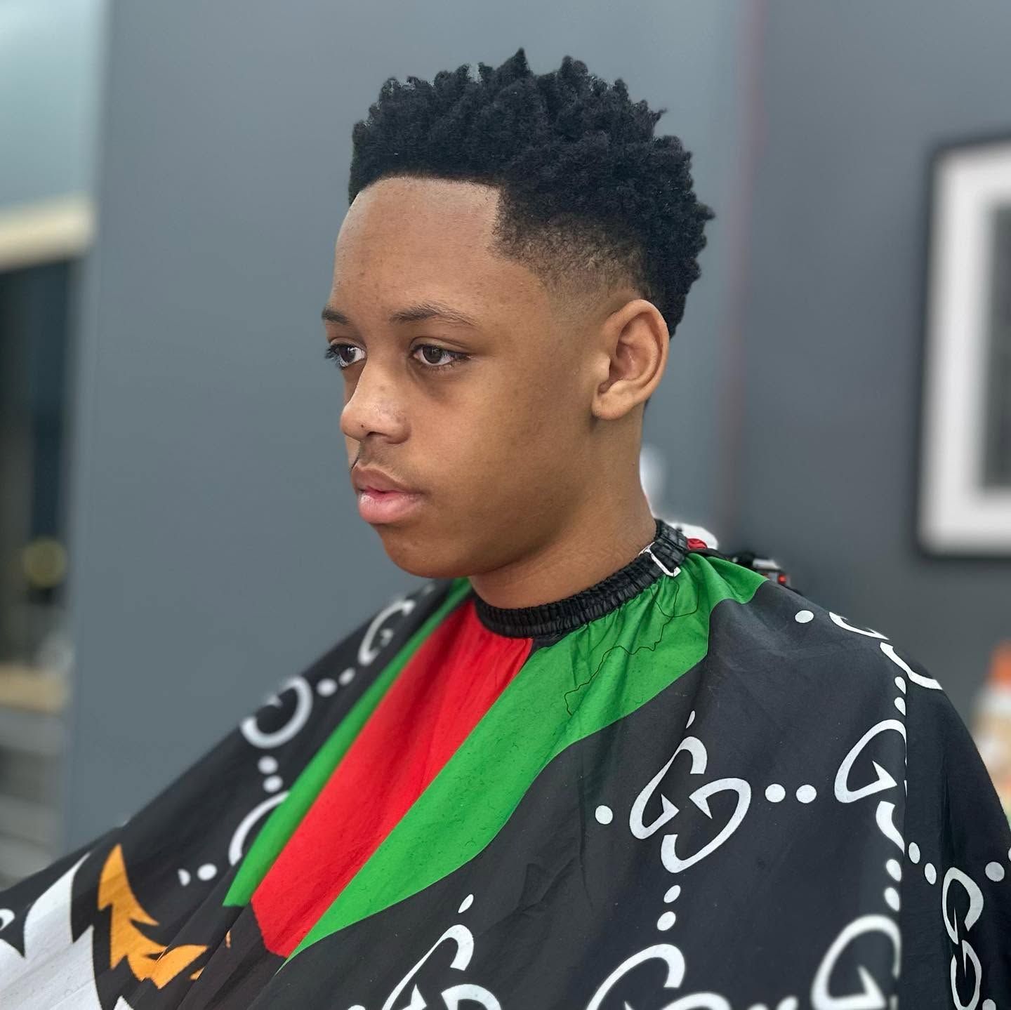 Person with a short haircut wearing a black, green, and red barber cape, seated in a salon.