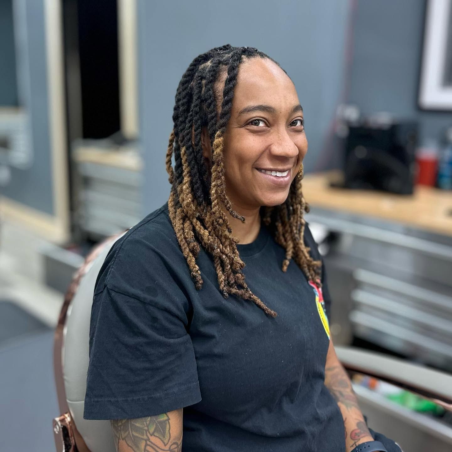Smiling person with braided hair seated in a salon chair, wearing a black T-shirt.