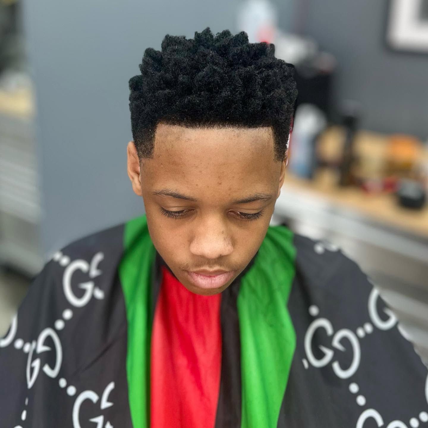 Person in a black, green, and red barber cape, looking down in a barbershop chair.