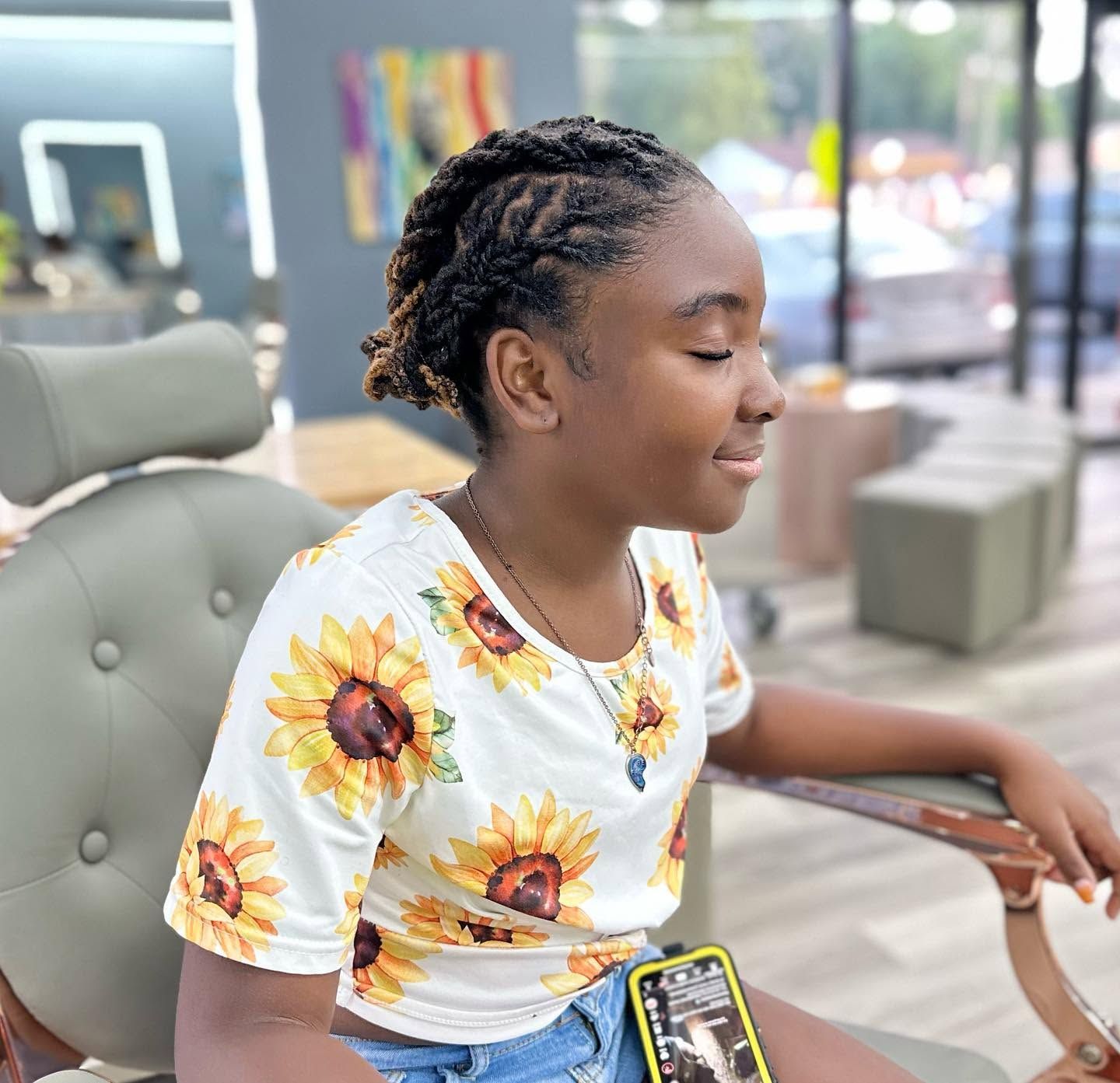 Person sitting in a salon chair, wearing a sunflower shirt, with braided hair and a phone on their lap.