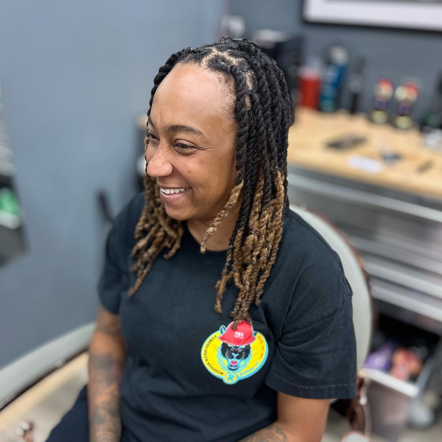 Smiling person with long braids in a black shirt, seated in a salon chair