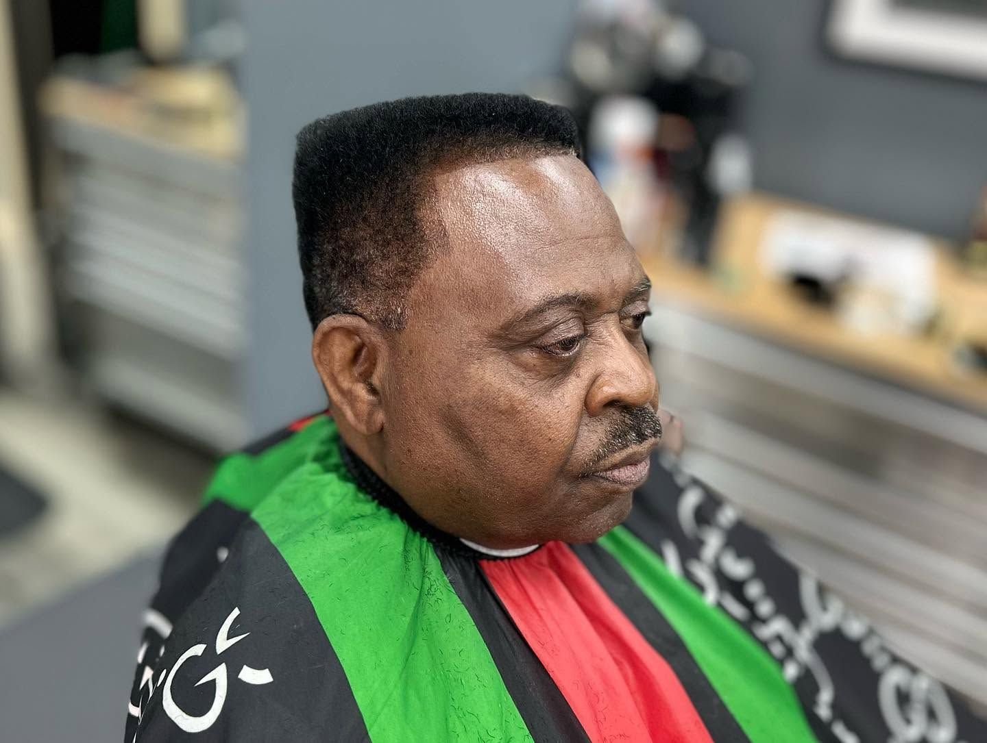 Man sitting in a barber chair wearing a black cape with green and red panels, in a salon setting
