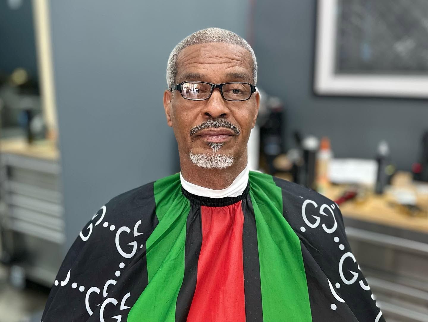 Man seated indoors wearing glasses and a black, green, and red patterned robe, looking at the camera.