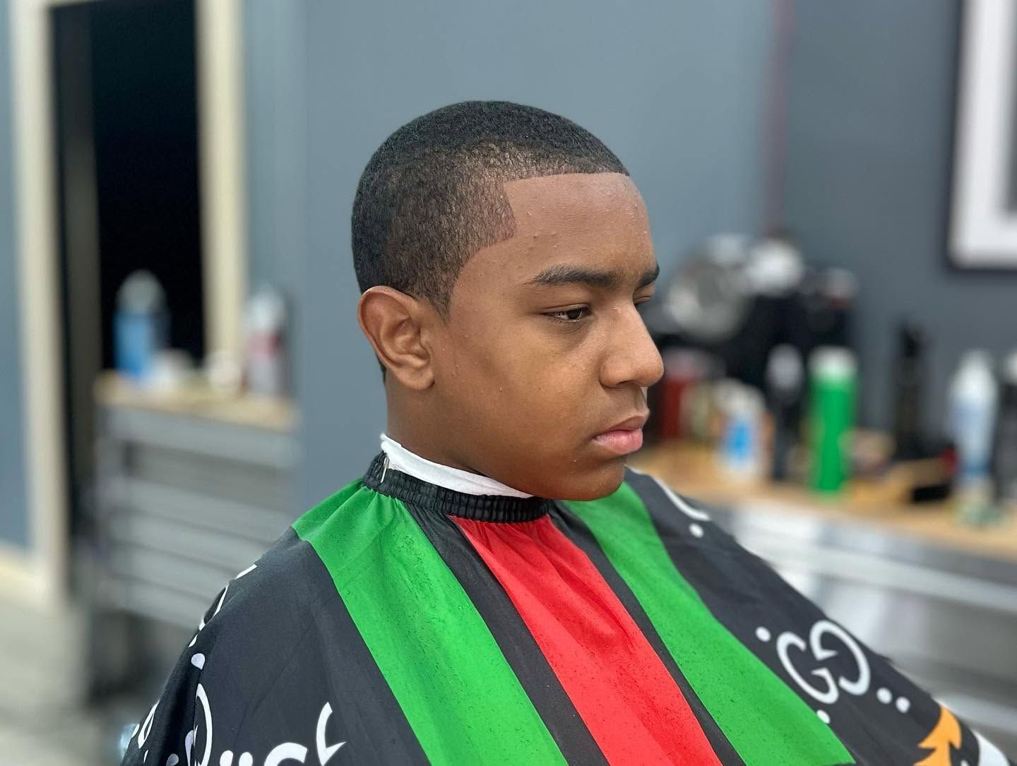 Boy in a barber cape with green, red, and black stripes, looking down in a barbershop.