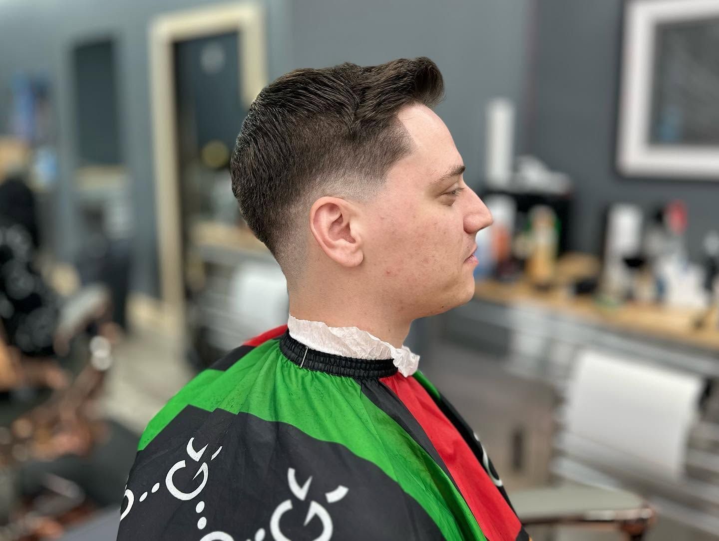 Man seated in a barber chair with a fresh fade haircut, wearing a green-and-black cape in a salon.